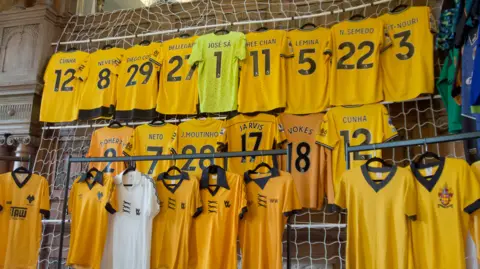 Twenty four Wolves shirts on display, hanging on three rows in a hotel suite. Twenty two are old gold, one is a green goalkeeper's jersey and one is a white away game shirt