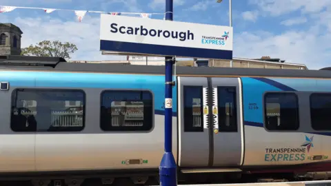 LDRS/Anttoni James Numminen A blue TransPennine Express train at the platform in Scarborough. A sign reading Scarborough is attached to a pole, bunting can be seen in the background.
