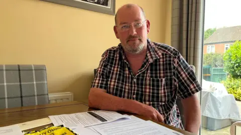 A man sat at a table wearing a checked shirt and glasses. He has short brown hair and a grey beard. On the table in front of him are piles of papers and a parking fine in bright yellow 