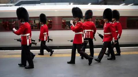 PA Media Seven guards walking alongside a white and red LNER train in full uniform. They are holding black, large guns.