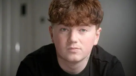 Close-up of Alex's face in the foreground, with short curly ginger hair and a dark top, looking towards the camera. A small neck tattoo is visible above the collar. The background is an indoor setting with a plain wall and a blurred door handle or fixture on the left. No bags, bins, or visible markers are present.