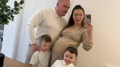 A selfie picture of a man, a woman and two young boys in front of a mirror. The wall and door behind them are white, there is a brown wooden table in front of them and there is a plant in the corner. The man and two boys all have short fair hair and are wearing white tops. The woman has long brown hair, is wearing a brown and white patterned dress, has a baby bump and is holding a phone on one hand with her other arm around the man.