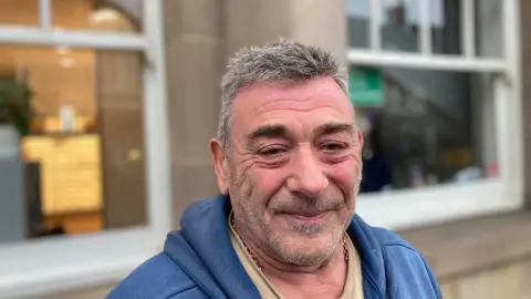 Laura Coffey/BBC A man smiles at the camera. He is wearing a blue hooded jumper and light coloured t-shirt. He has a gold chain round his neck. He has grey hair and stubble on his face.
