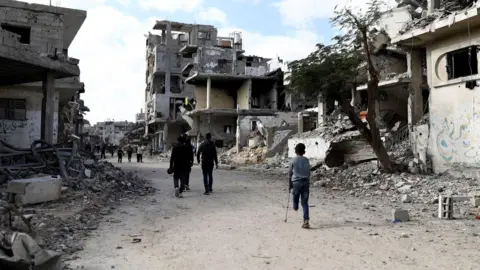 Reuters People, including a child on a crutch who is missing the lower half of his left leg, walk past the dusty gray rubble of homes and buildings destroyed during the war in Al-Bureij, central Gaza.