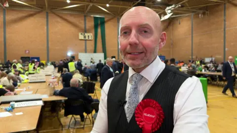 A man in a tie, white shirt and black waistcoat is wearing a red Labour rosette. Many people are inside behind him sitting at tables.