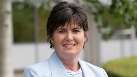 Solihull Council A woman with short dark hair smiles at the camera. She is wearing a blue-and-white-striped top under a pale blue blazer.