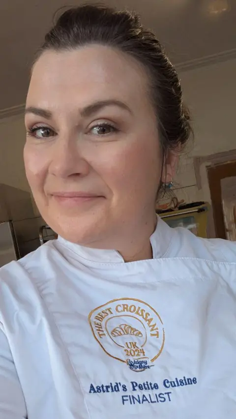 Astrid Roussel Astrid Roussel wears her white chef's uniform engraved with the logo of 2024's Best Croissant Competition. Astrid is a woman with brown hair which she wears up. She is smiling.