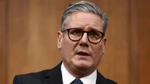 PA Media Sir Keir Starmer, a grey haired man with dark-rimmed glasses, photographed while speaking. He is wearing a dark suit and white shirt, in front of wooden wall. He is visible from the shoulders up. 