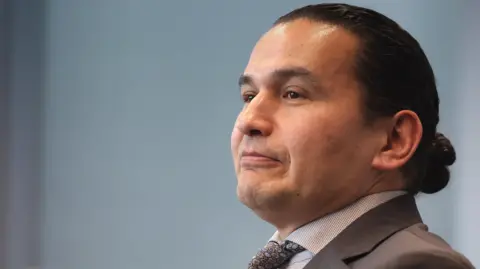Manitoba Premier Wab Kinew is seen in three-quarter profile with a neutral expression on his face. There is a pale blue non-descript background and he is wearing a grey suit and tie. 