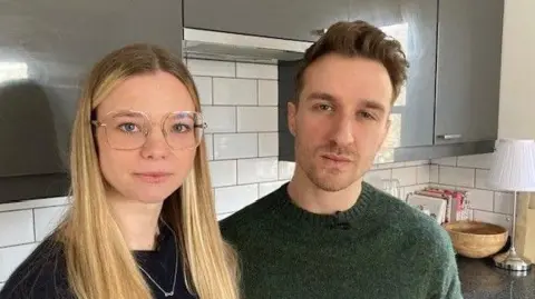 Jonah Kavanagh and Elle Van Pertersen stand in the kitchen in their flat with cupboards behind them.