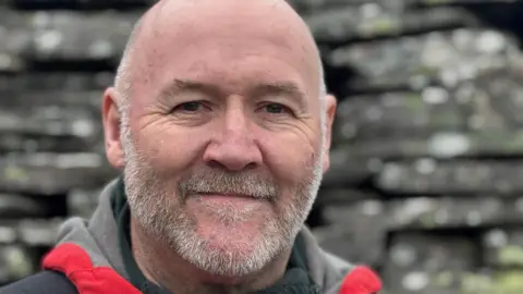 A middle-aged man with a short grey beard is looking into the camera. He is mostly bald. He is wearing an outdoor coat, with the red trim of the coat showing. Behind him, out of focus is a wall made of grey slate slabs.