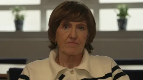 Woman with short, cropped brown hair, looking sternly at the camera. She wears a white jumper with horizontal black stripes on it.