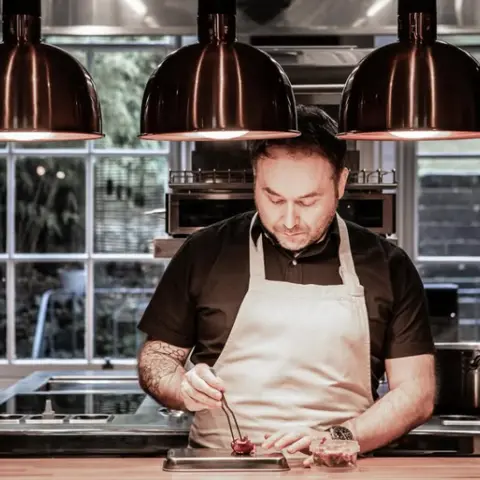 Lyla Stuart Ralston works at a bench in the kitchen, with tweezers placing the finishing touches to a food creation. He wears a cream apron. The lighting is moody with three pendant lights above, and windows behind him looking out into a garden area.