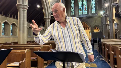Graham Harvey stands at a lectern in a church, as he conducts. He wears white trousers and a striped shirt and behind him are pews and a number of stained glass windows.