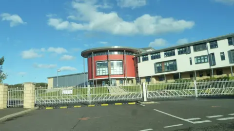 A modern school building with a round red section and the rest in white behind gates