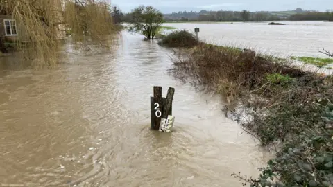 A river has burst its bank near a house in the countryside. The water is brown and covering most of the area in the picture.