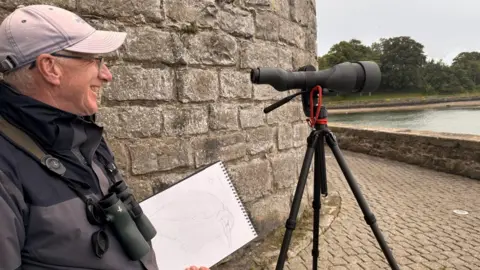 Richard Partis Richard Partis looks out at water smiling. He holds a sketchbook and stands behind his camera lense. He wears a grey jacket, black binoculars and a grey cap. 