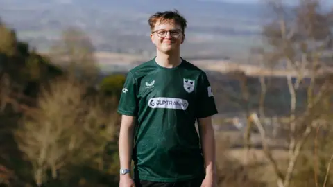 Dreams smoothly in stores and trees behind him, smiling in the camera. He wore glasses and top of green balls.