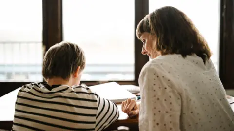 Getty/Maskot The back of a child wearing a stripey T-shirt sat at a table with books in front of him, with a woman sat next to him wearing a white blouse, turning slightly so a little of her side profile can be seen.