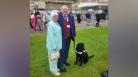 John Hardy with his arm around his wife's shoulders. They are both in suits standing in the garden of Buckingham Palace. John is holding Sidney, a black Labrador, on a lead.