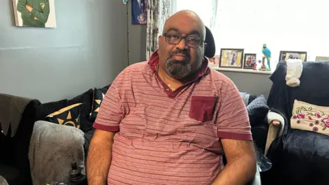 Claudia Sermbezis/BBC Thirugnanam 'Thiru' Sureshan is sitting in a chair in a living room