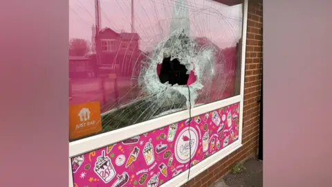 Catling Bakery's smashed window. A large hole has been made in the middle of the pane of glass and cracks have spread across the rest of the window. A pink banner below the window shows cartoons of cakes and milkshakes.