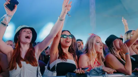 Sundown Festival 2024/jakhoward The front row of an outdoor gig, with five young women, all with long hair, raising their arms, under a blue sky