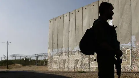 An Israeli soldier on patrol beside a protective security barrier decorated with graffiti that reads "Path for Peace" in Hebrew near the border fence with Gaza Strip, left, in the moshav Netiv HaAsara.
