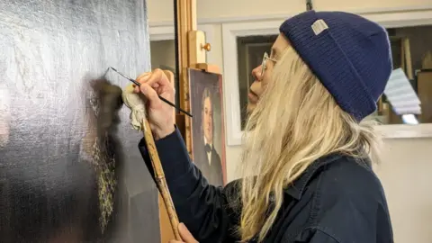 A woman with long blonde hair touches up a dark portrait on an easel with a very thin paintbrush. She wears glasses, a blue beanie hat and a dark blue shirt.