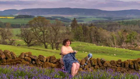 Barwood/Aynsley The image shows a woman sitting on a stone wall, with fields and hills behind her. She is naked apart from some strategically-placed knitting covering part of her body 