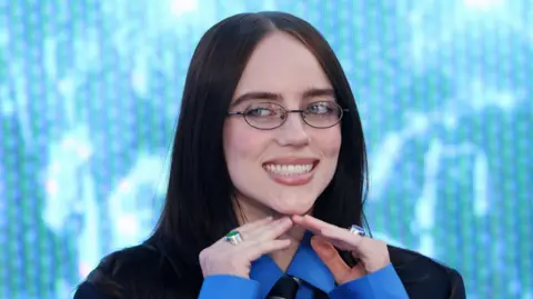 EPA/Shutterstock Billie Eilish on the blue carpet at the London premiere of her 3D concert film, Hit Me Hard And Soft: The Tour. She wears a blue shirt with a navy blue striped tie, and a dark coloured blazer. Her long brown hair sits on her shoulders, and she wears glasses. 
