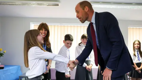 PA Media William in a class room bending to shake hands with a pupil, Ruby, who is smiling up a him.