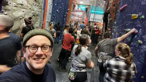 Simon Mutter A man stands in a crowded room where people are climbing up walls. 