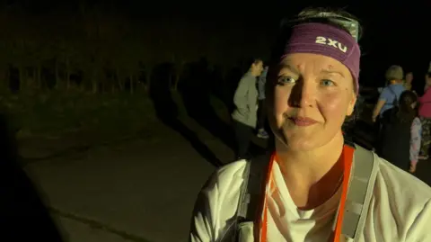 Ollie Rawlinson/BBC Nicky looking to the left of the camera pursing her lips.. She has her hair tied back and she is wearing a purple headband. She is also wearing a white top and grey running vest with the same red medal as Donna around her neck.
