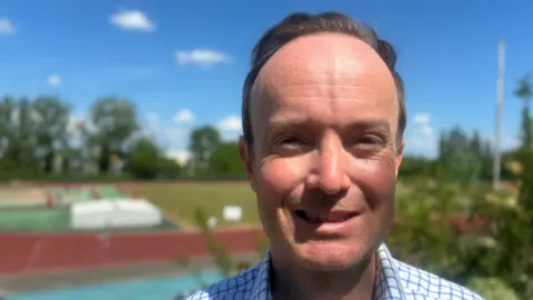 Matt Precey/BBC A man smiles at the camera in a head and shoulders image. He has brown hair and he is squinting slightly in bright sunshine. He is wearing a white shirt with a blue grid pattern on it.