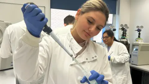 A woman in a white medical coat holding a pipette and a sample pot