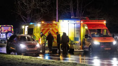 Ambulance vehicles and a mountain rescue vehicle parked on a road. A car is parked next to the emergency vehicles. It is dark. A group of people are stood next to the vehicles. 