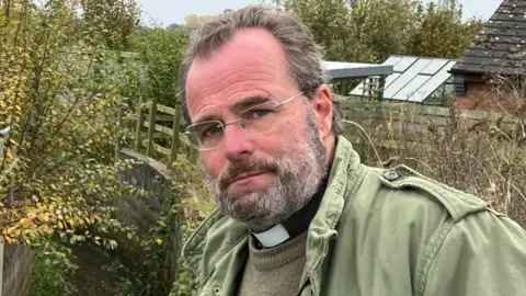 A man with dark hair, greying beard, glasses and wearing a vicar's collar under a green jacket and brown jumper. He is standing by some fencing and trees.