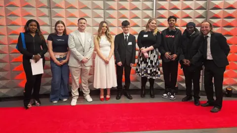 A line up of college students dressed in black tie posing for a picture against a grey 3D backdrop, standing on a red carpet.