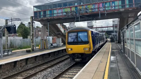 Photograph of a train at Macclesfield Station. The image shows a train, two platforms and a footbridge.