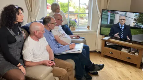 Show five people, including two women on either side of three men, sitting on a settee watching television