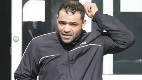 Lucian Sorenau wearing a black tracksuit top and scratching his head. He is looking at the camera.