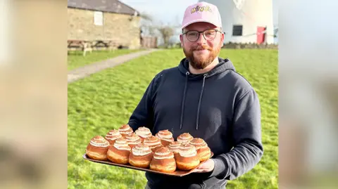 Richard Holt wearing a pink cap hat, a dark hoodie and glasses. He is holding a tray of doughnuts with cream topping and chocolate dusting. 