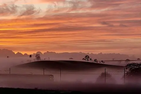 Sharon Macpherson A train going through low-lying mist at sunset, with fields and trees in the distance.