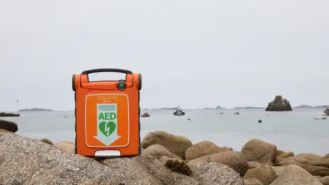BHF/Inga Drazniece A orange defibrillator on top of a rock by the sea. It has the letters 'AED' in green at the centre, with a green heart with a lightening bolt in the middle. The rocks are grey and brown. 