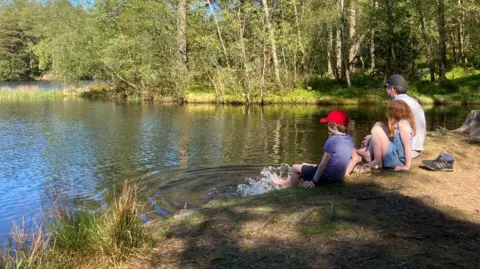 Patrick Charnley Patrick sitting and looking out across the Lake District with his children, who have their toes dipped in the water 
