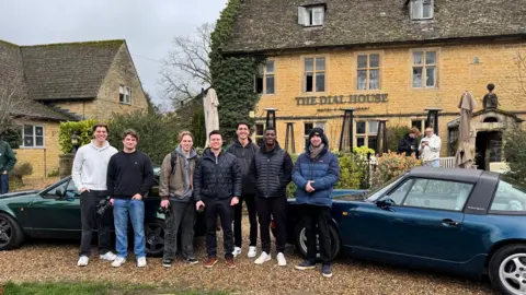 A group of seven young men stand in front of two older convertible cars, one is navy and the other is dark green. The cars are parked outside a Cotswold stone inn called The Dial House on a cloudy day.