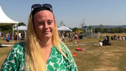A young mum with long blonde hair smiles as she stands in a scorched field on a hot summer day. Behind her, children are playing games, with outdoor activities like swingball, and gazebo-style tents. She is wearing a floral, green and white tunic-style top, a black headband, and sunglasses on top of her head. She has a nose ring.