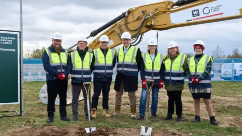 WELLCOME GENOME CAMPUS A line of four men and three women, standing next to each other and smiling at the camera. They are wearing hi-vis jackets, hard hats and gloves. In front of them, there are two spades in a patch of brown earth. Behind them is a crane. 