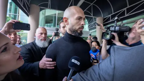 Reuters Andrew Tate, bearded and wearing a black jacket, surrounded by pressure outside the airport station